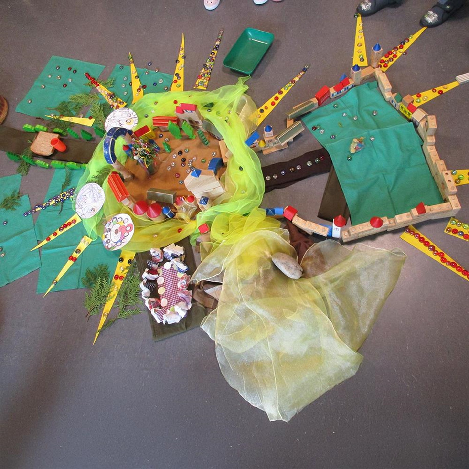 Das Bild zeigt eine fantasievolle Bodenlandschaft, gestaltet mit bunten Tüchern, Holzklötzen, Spielmaterialien und gebastelten Sonnenstrahlen aus Papier. Kleine Figuren, glänzende Steine und bemalte Scheiben sind in die Szene eingebaut – vermutlich ein gemeinsames Projekt von Kindern, das eine symbolische Welt oder Geschichte darstellt. Die kreative Anordnung und die verwendeten Materialien deuten auf ein pädagogisches Konzeptspiel oder eine Gruppenarbeit im Kindergarten hin.
