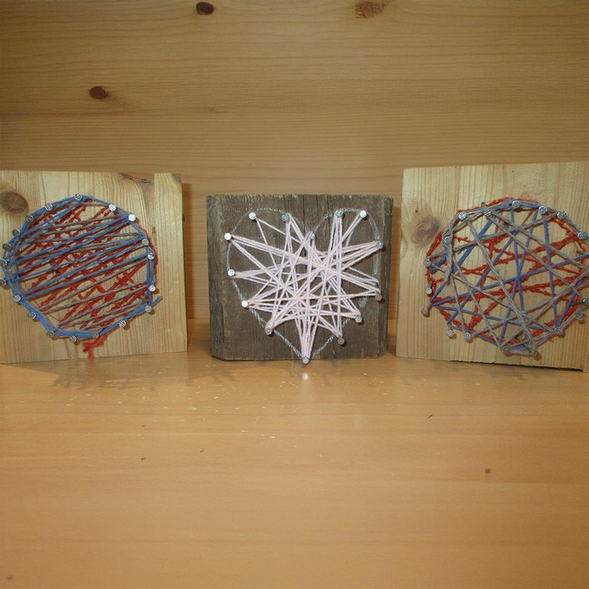 Das Bild zeigt drei selbstgemachte Fadenbilder auf kleinen Holzplatten. Zwei Motive sind kreisförmig mit überkreuzten roten und blauen Fäden gestaltet, das mittlere Bild zeigt ein Herz aus rosafarbener Wolle. Die Nägel, an denen die Fäden befestigt sind, bilden die Konturen – typisch für Kinderbasteleien oder kreative Projekte im Kindergarten oder Grundschulbereich.