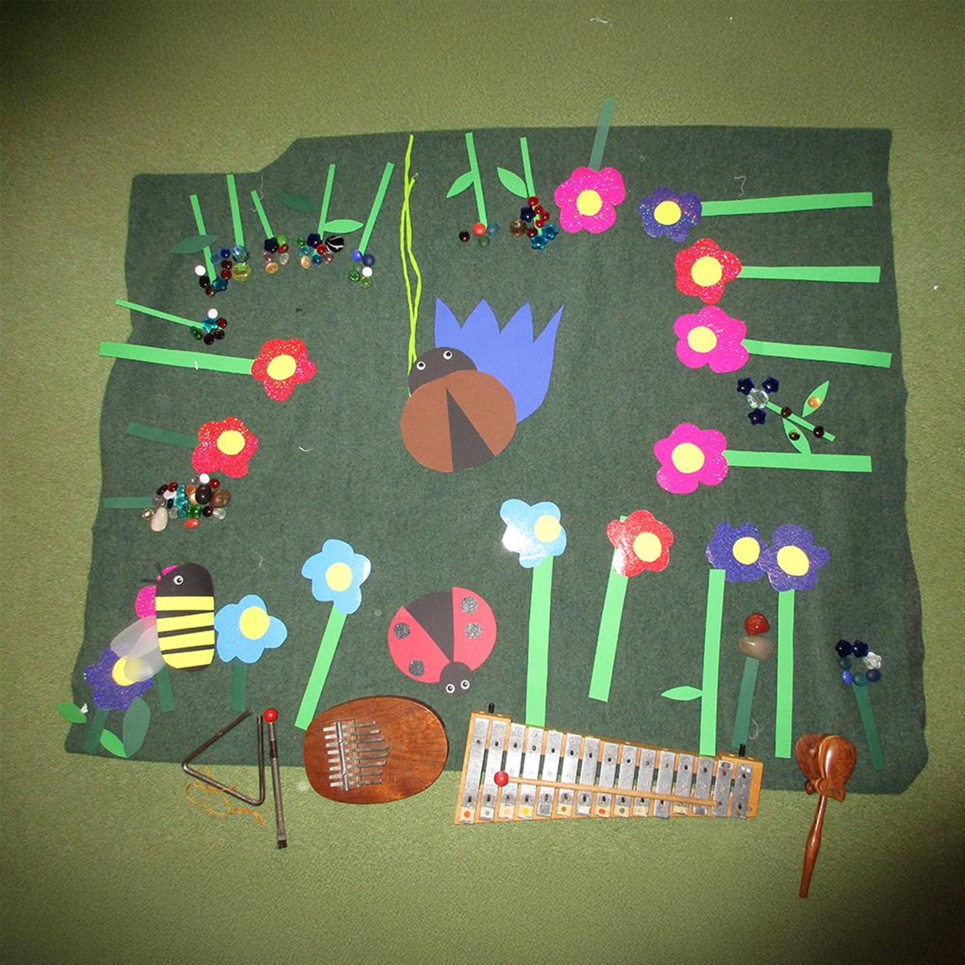 Das Bild zeigt ein kreativ gestaltetes Frühlingsbild auf grünem Filzuntergrund mit bunten Papierblumen, Marienkäfer, Biene und einer aufgehängten Spinne. Um die Blumen herum sind dekorative Perlen und Glitzersteine angebracht. Am unteren Bildrand liegen verschiedene Musikinstrumente – ein Xylophon, eine Kalimba, ein Triangel und ein Maracas – was auf ein musikalisches Angebot oder eine thematische Einheit in der Kita hinweist.