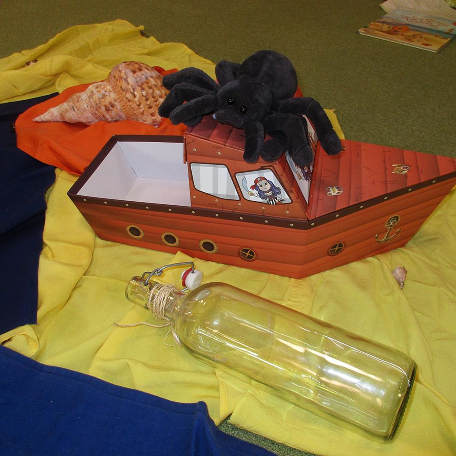 Das Bild zeigt eine fantasievolle Spielszene: Ein Pappschiff mit Piratenmotiv steht auf bunten Tüchern, die Wasser und Strand symbolisieren. Auf dem Schiff sitzt eine große Plüschspinne, daneben liegen eine große Muschel und eine leere Flaschenpost aus Glas. Im Hintergrund sind Kinderbücher zu sehen – vermutlich Teil eines thematischen Projekts oder Rollenspiels in einer Kita oder Vorschulgruppe.