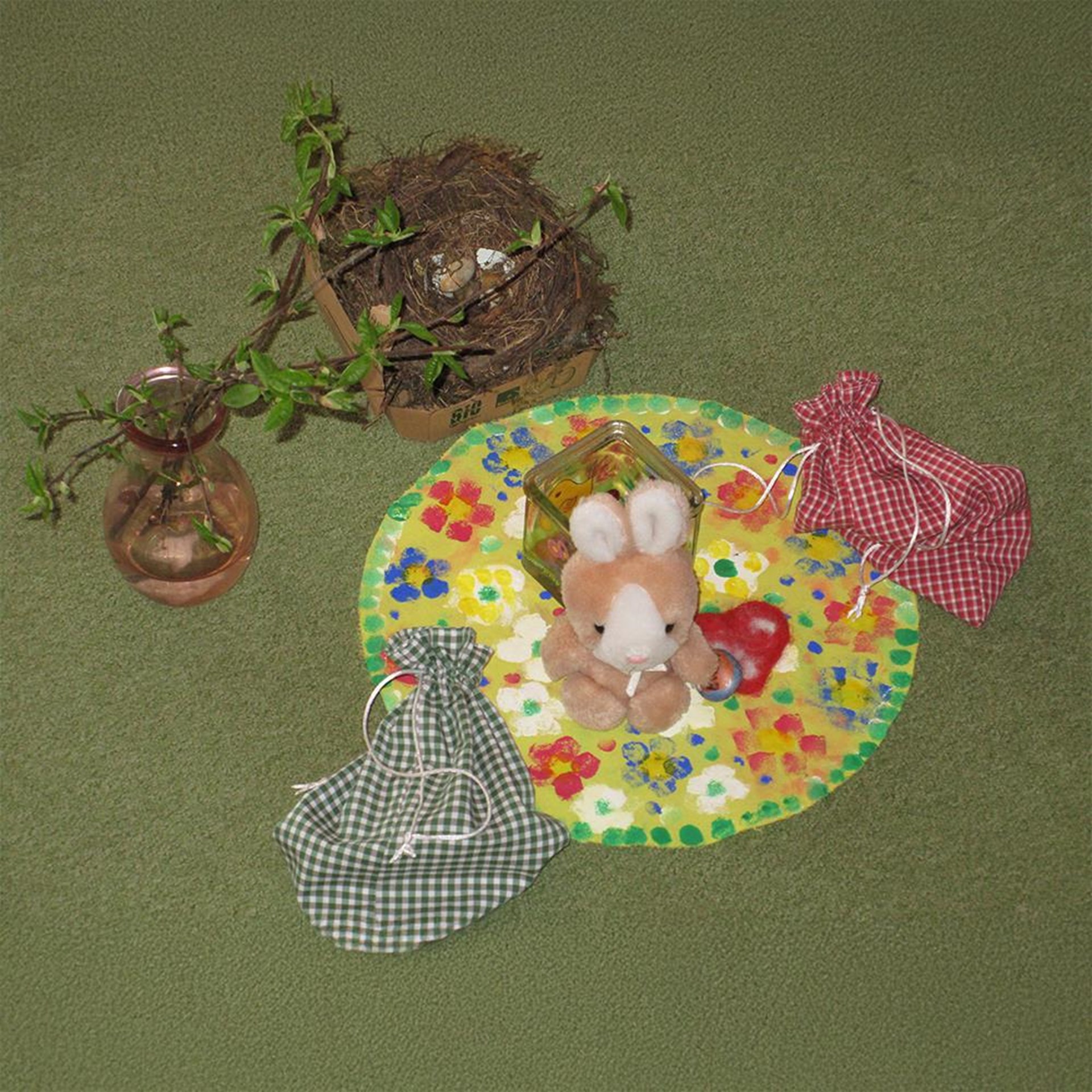 Das Bild zeigt eine frühlingshafte oder österliche Deko-Szene auf grünem Teppichboden: In der Mitte sitzt ein Plüschhase auf einem bunt bemalten, blumenförmigen Papierkreis, daneben ein Herz und ein Glas mit Motiv. Zwei kleine Stoffbeutel in Rot und Grün liegen daneben, und im Hintergrund steht ein Glas mit frischem Zweig sowie ein Nest mit Vogeleiern in einer Obstkiste. Die Gestaltung wirkt liebevoll und kindgerecht – vermutlich Teil einer pädagogischen Aktivität im Kindergarten.